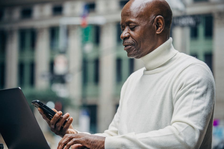 Side view of concentrated African American businessman in casual clothes browsing smartphone while working on laptop on city street