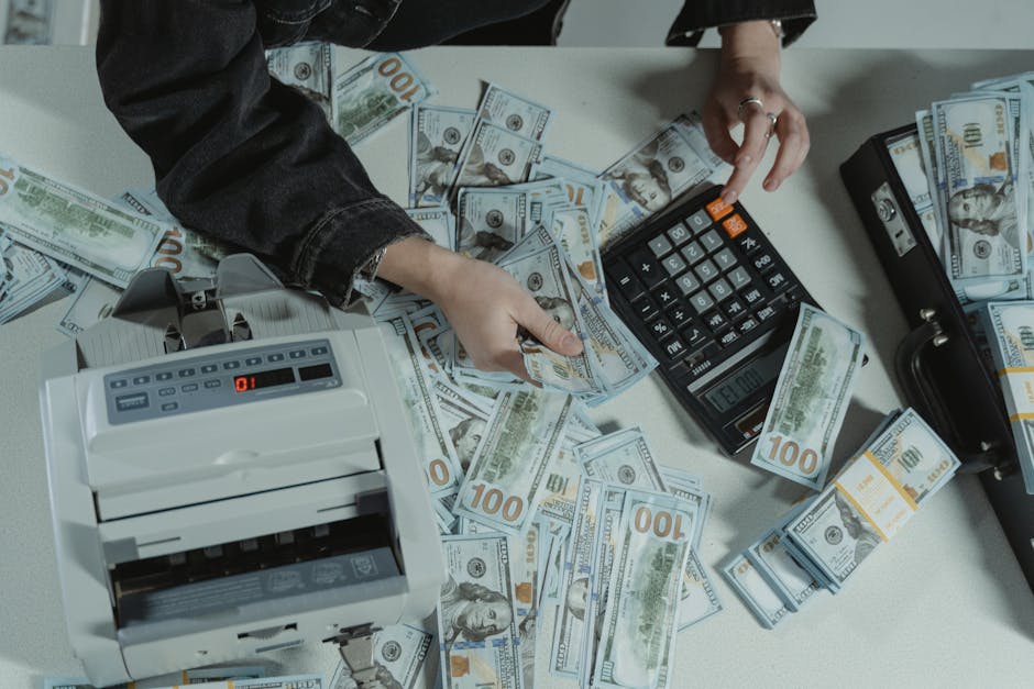 Hands counting 100 dollar bills using a calculator and money counter on a table.