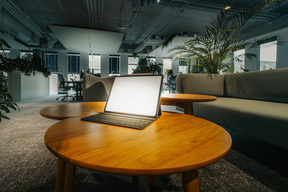 Empty office lounge featuring a glowing laptop on a wooden table, surrounded by greenery.
