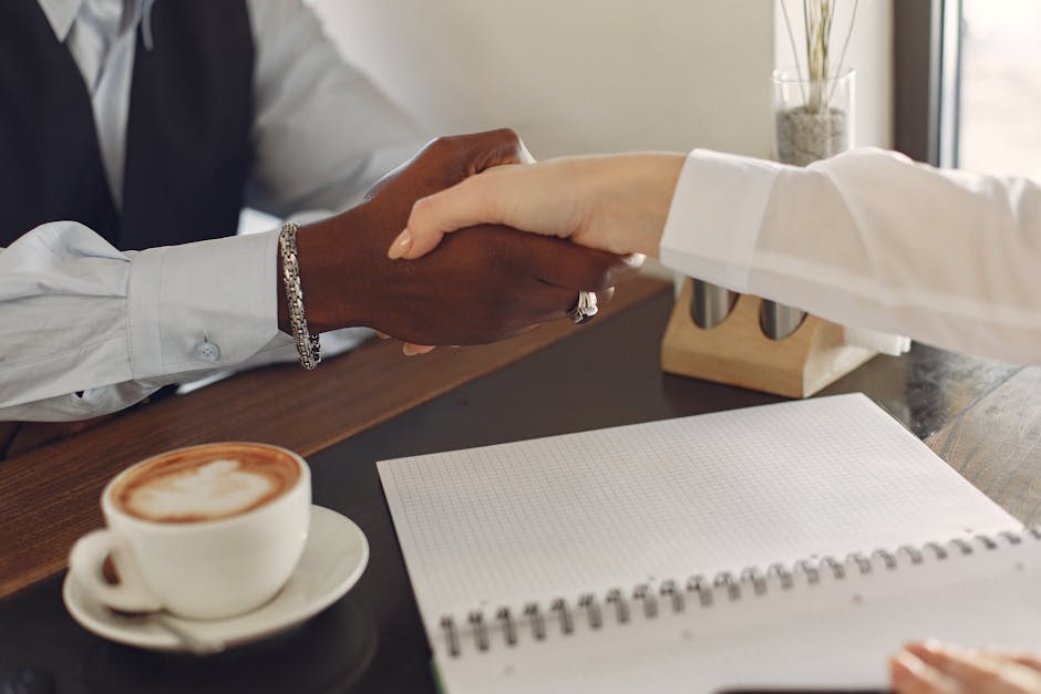 Two professionals shaking hands at a desk with a coffee cup and notebook, symbolizing business agreement.