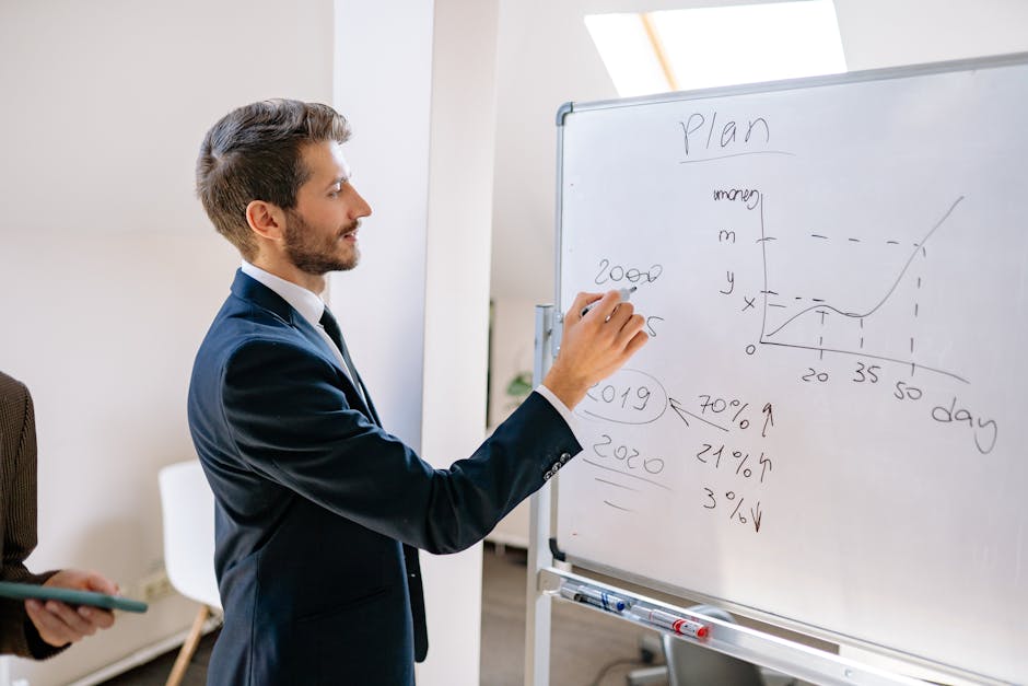 A businessman in a suit writes financial data on a whiteboard during an office planning session.