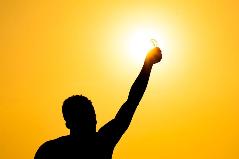 From below of silhouette of faceless male raising hand with light bulb against bright sundown sky