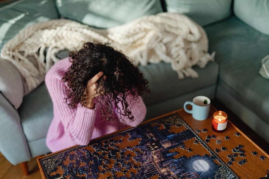 A woman in a pink sweater feels frustrated while solving a jigsaw puzzle indoors, surrounded by cozy decor.